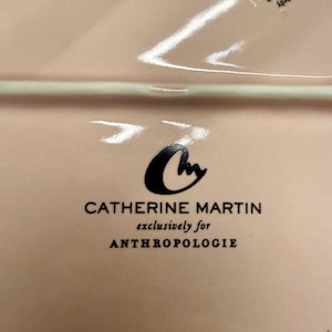 Op de afbeelding: Een lichtroze rechthoekige keramische plaat met het Catherine Martin-logo en de tekst "exclusively for ANTHROPOLOGIE". De plaat heeft een glanzende afwerking en is nieuw in de verpakking.