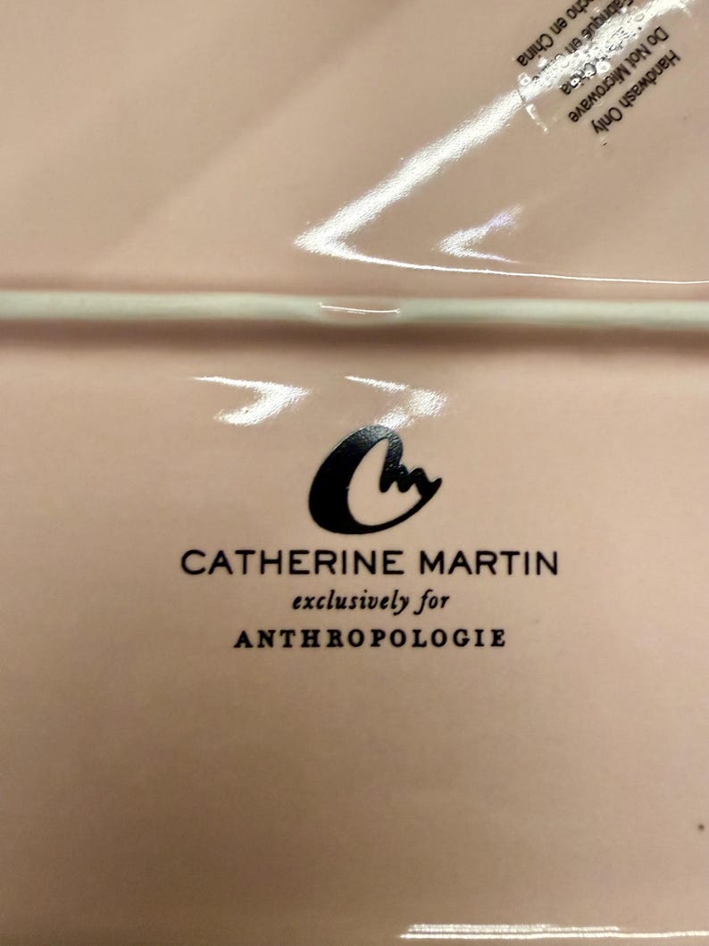 Op de afbeelding: Een lichtroze rechthoekige keramische plaat met het Catherine Martin-logo en de tekst "exclusively for ANTHROPOLOGIE". De plaat heeft een glanzende afwerking en is nieuw in de verpakking.
