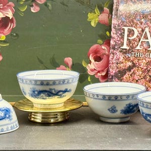 Könnte beinhalten: Eine Sammlung weiß-blauer Porzellanschalen mit Drachenmotiven. Eine Schale ist umgedreht. Die Schalen stehen auf einem goldenen Ständer. Im Hintergrund ein Buch mit dem Wort "PARIS".