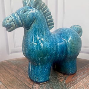 Puede incluir: Una figura de caballo de cerámica con un esmalte brillante en tonos azul profundo y verde azulado con un acabado texturizado. La escultura representa un caballo estilizado en posición de pie con una crin prominente, brida definida y cuerpo redondeado.