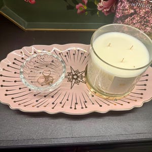 Op de afbeelding: Een roze decoratief dienblad met een gouden rand en een sterrenpatroon bevat een heldere glazen kom met ringen en een grote kaars in een glazen pot. De kaars heeft een gouden voet en meerdere lonten.