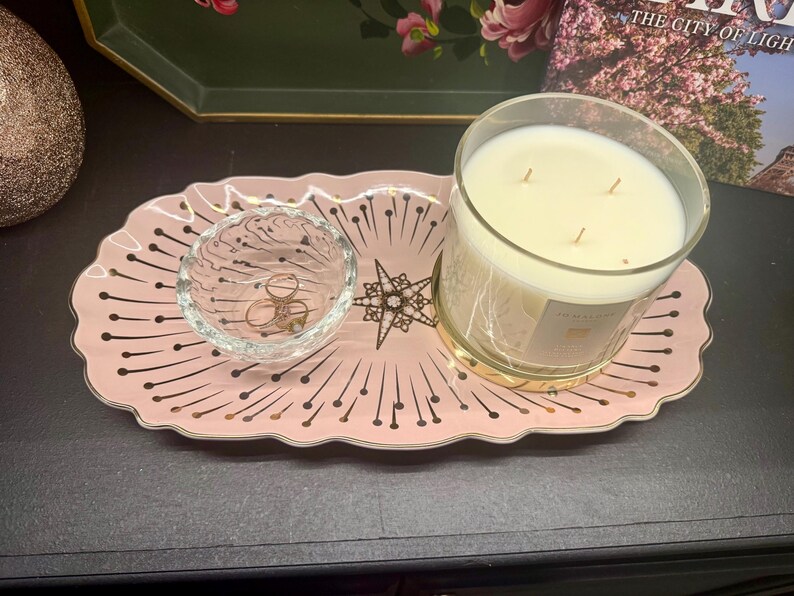 Op de afbeelding: Een roze decoratief dienblad met een gouden rand en een sterrenpatroon bevat een heldere glazen kom met ringen en een grote kaars in een glazen pot. De kaars heeft een gouden voet en meerdere lonten.