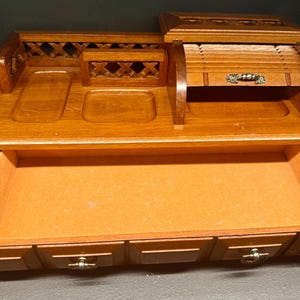 May include: A vintage wooden desk organiser in a warm, medium-brown finish. It features a roll-top compartment, small drawers, and open compartments. The organiser has a felt-lined drawer and decorative brass hardware. A classic piece for home or office.