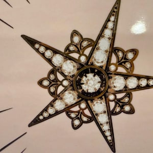 Op de afbeelding: Close-up van een decoratief sterrenburst-ontwerp. De ster heeft vijf punten met heldere, ronde versieringen en een donker bronzen metalen frame. De achtergrond is lichtroze met zwarte lijnen die uitstralen van de ster.