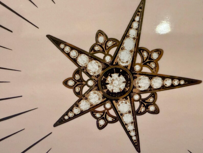 Op de afbeelding: Close-up van een decoratief sterrenburst-ontwerp. De ster heeft vijf punten met heldere, ronde versieringen en een donker bronzen metalen frame. De achtergrond is lichtroze met zwarte lijnen die uitstralen van de ster.
