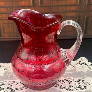 May include: A ruby red glass pitcher with a clear, textured handle. The pitcher has a wide spout and a rounded body with a repeating floral pattern. The pitcher is sitting on a white lace tablecloth.