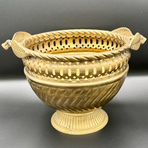 Könnte beinhalten: Eine Vintage-Messingschale mit dekorativem Design. Die Schale hat einen durchbrochenen Rand, eine seilartige Bordüre und blattförmige Griffe. Die Schale hat einen Sockel und eine strukturierte Außenseite. Die Gesamtfarbe ist ein warmes, antikes Gold.