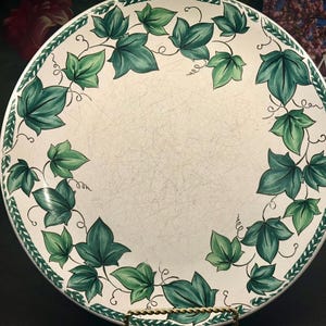 Op de afbeelding: Ronde, crèmekleurige decoratieve schaal met een groen klimopbladmotief langs de rand. De bladeren zijn geschilderd in groentinten, met een getextureerd oppervlak. De schaal wordt gepresenteerd op een goudkleurige standaard.