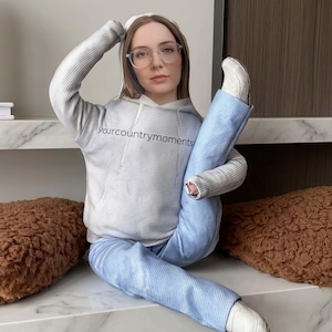 Puede incluir: Una sudadera con capucha gris claro con el texto "yourcountrymoments" en negro, combinada con vaqueros azul claro. La figura está sentada, con una pierna extendida hacia arriba. La figura lleva gafas y zapatos blancos.