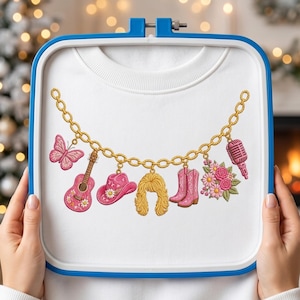 May include: A white sweatshirt featuring an embroidered gold chain design with hanging charms including a pink butterfly, a pink acoustic guitar, a pink cowboy hat, blonde hair, pink cowboy boots, a bouquet of pink flowers, and a pink microphone.