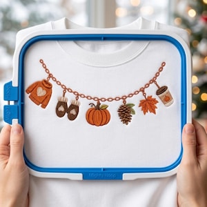 May include: A white sweatshirt secured in a blue embroidery hoop featuring an embroidered chain design with hanging charms including an orange sweater, brown mittens, a pumpkin, a pinecone, an autumn leaf, and a coffee cup with a pumpkin icon.