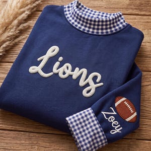 May include: A navy blue long-sleeve shirt featuring a mock neck collar with a blue and white gingham pattern. The word Lions is embroidered in white script on the chest. The sleeve features an embroidered football and the name Zoey in white.