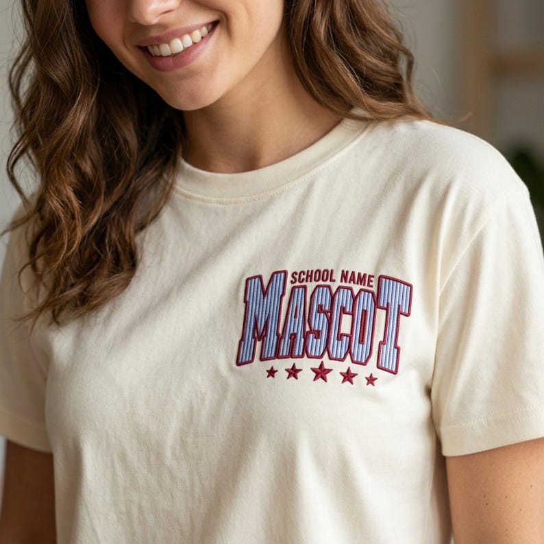 May include: A cream-colored short-sleeve t-shirt featuring an embroidered design on the left chest. The embroidery includes the text "SCHOOL NAME" above the word "MASCOT" in blue and white striped letters with a dark red outline and five stars.