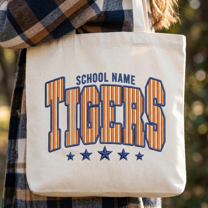 May include: A cream-colored canvas tote bag featuring the text SCHOOL NAME in dark blue above the word TIGERS in large, orange and white striped varsity-style lettering with a dark blue outline, accented by five dark blue stars below.