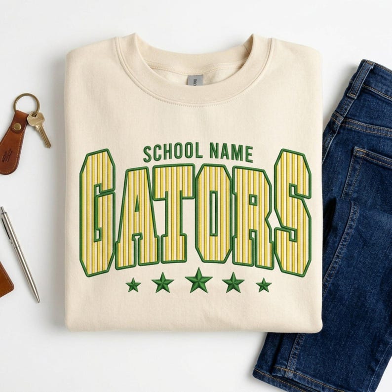 May include: A cream-colored crewneck sweatshirt featuring the text SCHOOL NAME in green above the word GATORS in large, yellow, vertically striped letters with green outlines, finished with five green stars centered below the text.