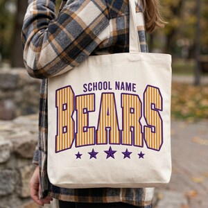 May include: A cream-colored canvas tote bag featuring the text SCHOOL NAME in dark purple letters above the word BEARS in large, orange and yellow striped block letters with a dark purple outline, finished with five purple stars below.