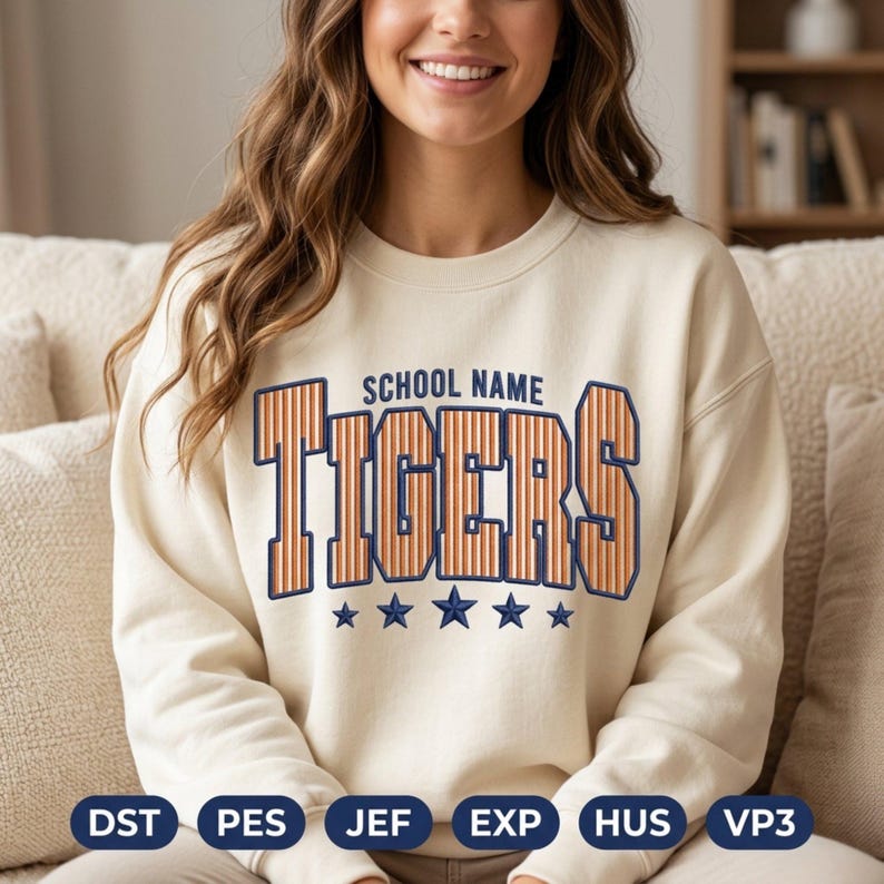 May include: A cream-colored crewneck sweatshirt featuring the text SCHOOL NAME in small dark blue letters above the word TIGERS in large, orange, vertically striped block letters with a dark blue outline, and five dark blue stars below.