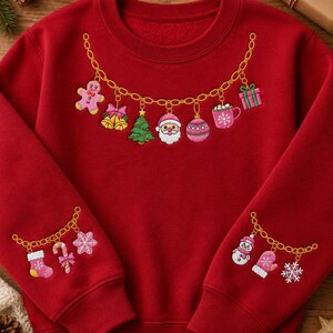 May include: A red crewneck sweatshirt featuring a gold chain graphic across the chest and sleeves. The chest chain displays charms of a gingerbread person, bells, a tree, Santa, an ornament, a mug, and a gift. Sleeves feature stocking, candy cane, snowflake, snowman, and mitten charms.
