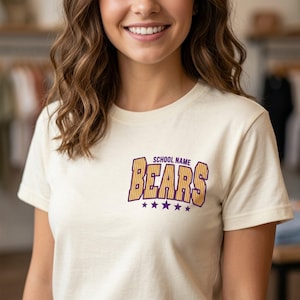 May include: A cream-colored short-sleeve t-shirt featuring a graphic on the chest. The graphic displays the text "SCHOOL NAME" in small purple letters above the word "BEARS" in large, yellow, striped block letters, with five purple stars below.