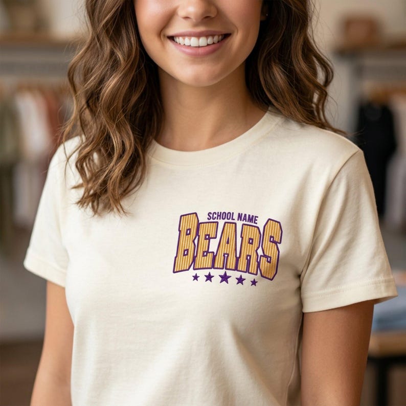 May include: A cream-colored short-sleeve t-shirt featuring a graphic on the chest. The graphic displays the text "SCHOOL NAME" in small purple letters above the word "BEARS" in large, yellow, striped block letters, with five purple stars below.