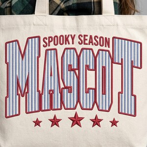 May include: A cream-colored canvas tote bag featuring the text SPOOKY SEASON MASCOT in large, block-style letters with a blue and white pinstripe pattern and red outlines. Below the text are five red embroidered stars arranged in a horizontal row.