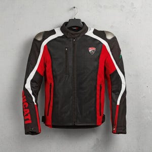 Pode incluir: Casaco de motociclista preto, vermelho e branco com a palavra "Ducati" estampada na manga. O casaco tem fecho de correr, um pequeno logótipo no peito e proteções de ombro prateadas. Está pendurado num cabide preto.