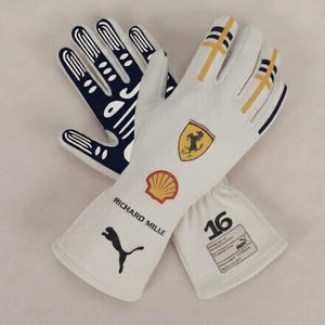 Peut inclure: Une paire de gants de course blancs arborant le logo Ferrari, un emblème Shell, le texte RICHARD MILLE, un logo Puma et le numéro 16. Les gants présentent des accents jaunes et noirs sur les doigts, avec une paume noire à motifs de lignes blanches.