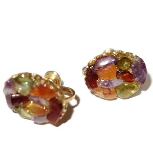 May include: A pair of gold-toned stud earrings. Each earring features a cluster of colorful gemstones in shades of red, orange, purple, and green. The gemstones are set in a gold-colored metal setting, creating a unique and eye-catching design.