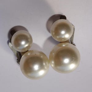 May include: A pair of clip-on earrings featuring large, cream-colored faux pearls. Each earring has two pearls of different sizes, attached to a silver-toned clip. The pearls have a glossy finish, reflecting light.
