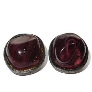 May include: Two vintage, round earrings. Each earring features a deep purple, domed glass stone with swirling patterns, set in a tarnished silver-tone metal bezel. The earrings are displayed against a white background.