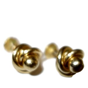 Puede incluir: Un par de gemelos dorados, cada uno con un diseño anudado. Los gemelos tienen un acabado brillante y pulido sobre un fondo blanco. Un accesorio clásico y elegante para ocasiones formales.