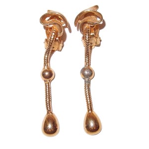 May include: A pair of gold-toned clip-on earrings. Each earring features a curved top, a textured, snake-like body, a spherical accent, and a teardrop-shaped bottom. The earrings are displayed against a white background.
