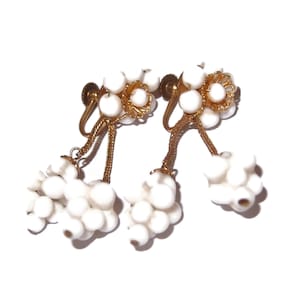 May include: A pair of vintage clip-on earrings featuring clusters of white beads and gold-tone metal accents. The earrings have a floral design at the top and a dangling cluster of beads, creating a delicate and elegant look.