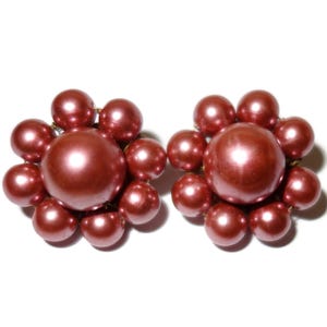 May include: A pair of vintage clip-on earrings featuring a cluster design. Each earring has a large, round, reddish-brown pearl surrounded by smaller pearls. The earrings are set against a white background, highlighting their color and shape.