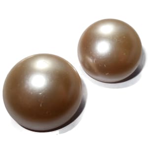 Puede incluir: Dos cuentas redondas, marrones, con aspecto de perla y acabado brillante. Las cuentas son de un color cálido y neutro, y parecen estar hechas de un material liso. Están sobre un fondo blanco.