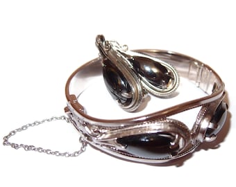 May include: A silver bracelet and matching earrings. The bracelet has three dark, oval stones and a safety chain. The earrings have a teardrop shape and dark stones. The jewellery set is displayed on a white background, showcasing the reflective silver.
