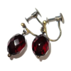 May include: A pair of vintage clip-on earrings featuring faceted, deep red gemstones. The earrings have a screw-back mechanism and are set in a gold-toned metal. The gemstones are oval-shaped and catch the light, creating a sparkling effect.
