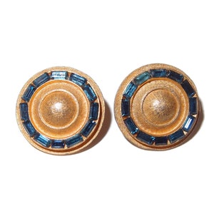 May include: A pair of gold tone earrings with a circular design. The earrings are decorated with a ring of blue rhinestones.