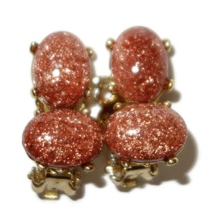 May include: Close-up of a pair of clip-on earrings. Each earring features four oval-shaped, reddish-brown stones with a glittery, speckled appearance. The stones are set in a gold-colored metal frame, creating a vintage jewelry aesthetic.