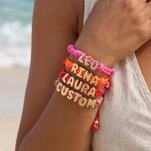 Könnte beinhalten: Vier gestapelte Armbänder aus leuchtend pinkfarbenen, orangen, roten und mehrfarbigen Kordeln. Jedes Armband ist mit goldenen Metallbuchstabenperlen versehen, die LEO, RINA, LAURA und CUSTOM buchstabieren. Verstellbare Knotenverschlüsse.