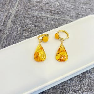 Op de afbeelding: Een paar goudkleurige oorbellen met druppelvormige barnsteenhangers. Elke oorbel heeft een kleine, ronde barnsteensteen aan de bovenkant, verbonden met een gouden ring. De barnsteenhangers zijn doorschijnend geel-oranje van kleur met zichtbare insluitsels.