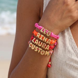Peut inclure: Un empilement de quatre bracelets en cordon tressé coloré, chacun orné de perles lettres en métal doré. Les bracelets forment les noms LEO, RINA, LAURA et le mot CUSTOM, avec chaque cordon dans une couleur vive différente, dont rose, orange et rouge.