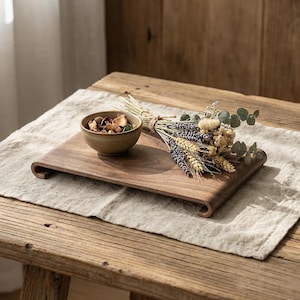 May include: A wooden serving tray with curved edges holds a small ceramic bowl and a bundle of dried flowers and wheat. The tray sits on a linen tablecloth atop a rustic wooden table, creating a natural, earthy aesthetic.