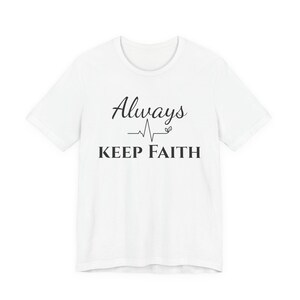 Op de afbeelding: Wit T-shirt met de tekst "Always Keep Faith" in zwarte letters en blokletters. Het ontwerp bevat een hartslaglijn en een klein blad. Het shirt heeft korte mouwen en een ronde hals.