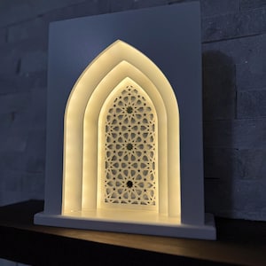 May include: A white, illuminated decorative lamp with an arched design. The lamp features a layered structure with a detailed star pattern in the center, emitting a warm, inviting glow. The lamp is placed on a dark surface against a textured wall.
