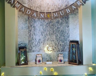 May include: A decorative Ramadan display featuring a banner that reads "RAMADAN MUBARAK". The display includes lanterns, decorative boxes, a crescent moon light, and a string of lights. The items are arranged on a shelf against a tiled wall.
