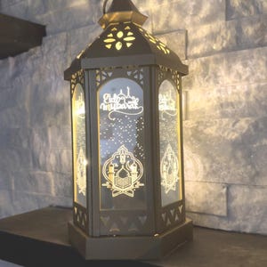 May include: A decorative, dark metal lantern with intricate cut-out designs and glass panels. The panels display the text "Eid Mubarak" and Islamic motifs. The lantern is illuminated from within, creating a warm glow.