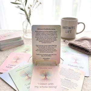 Puede incluir: Un juego de 36 tarjetas de afirmación titulado The Midwifery Journey, con ilustraciones de árboles en tonos pastel y texto. Una tarjeta se muestra en un soporte dorado, rodeada de otras tarjetas, una taza con Midwife Life y un florero de cristal.