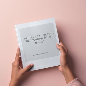 Peut inclure: Un livre blanc intitulé "MENTAL LOAD RESET - Das Entlastungs-Set für Mamas" est tenu par deux mains sur un fond rose. Le livre a un carré gris au centre avec le titre écrit en script noir. Le bas du livre porte le texte "@xmentalloadkicktes".