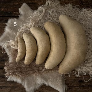 May include: A set of five crescent-shaped pillows in graduated sizes, arranged in a row from smallest to largest. Each pillow is made of a textured, light beige fabric. The number 5 is visible in the upper left corner of the image.